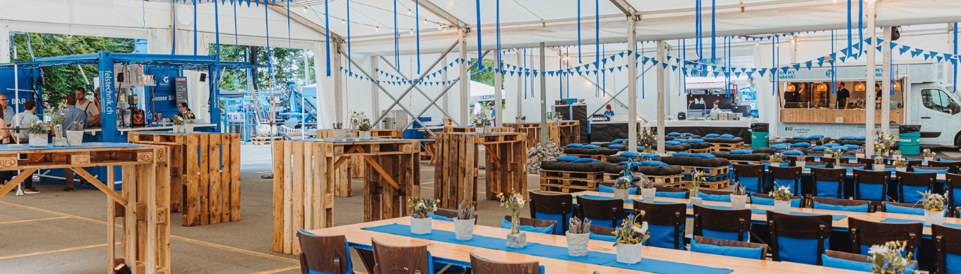 Festlich dekoriertes Zelt mit Tischen, Stehtischen, Sofas in der Firmenfarbe blau. 